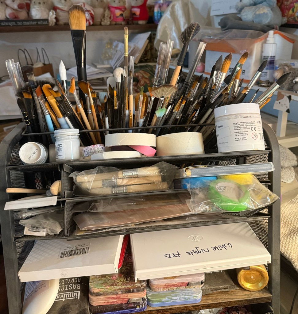 A cluttered artist's cart filled with various paintbrushes, jars, tapes, and art supplies organized in compartments.